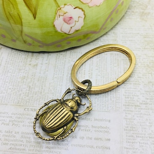 May include: A brass key chain featuring a detailed beetle charm. The beetle is a dark gold colour and has a textured surface. The key chain is attached to a brass oval ring.