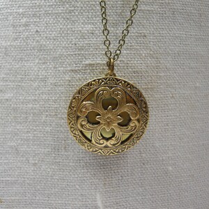 May include: A gold-colored round pendant necklace with an ornate floral design. The pendant is suspended from a delicate chain. The pendant features a detailed, embossed pattern with a central flower and surrounding scrollwork.