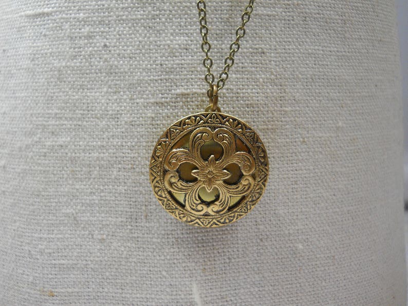May include: A gold-colored round pendant necklace with an ornate floral design. The pendant is suspended from a delicate chain. The pendant features a detailed, embossed pattern with a central flower and surrounding scrollwork.