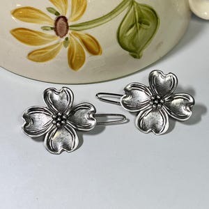 May include: Two silver-toned flower-shaped hair clips. Each clip features four petals and a cluster of small beads at the center. The clips are set against a light background, with a floral-decorated ceramic mug in the background.