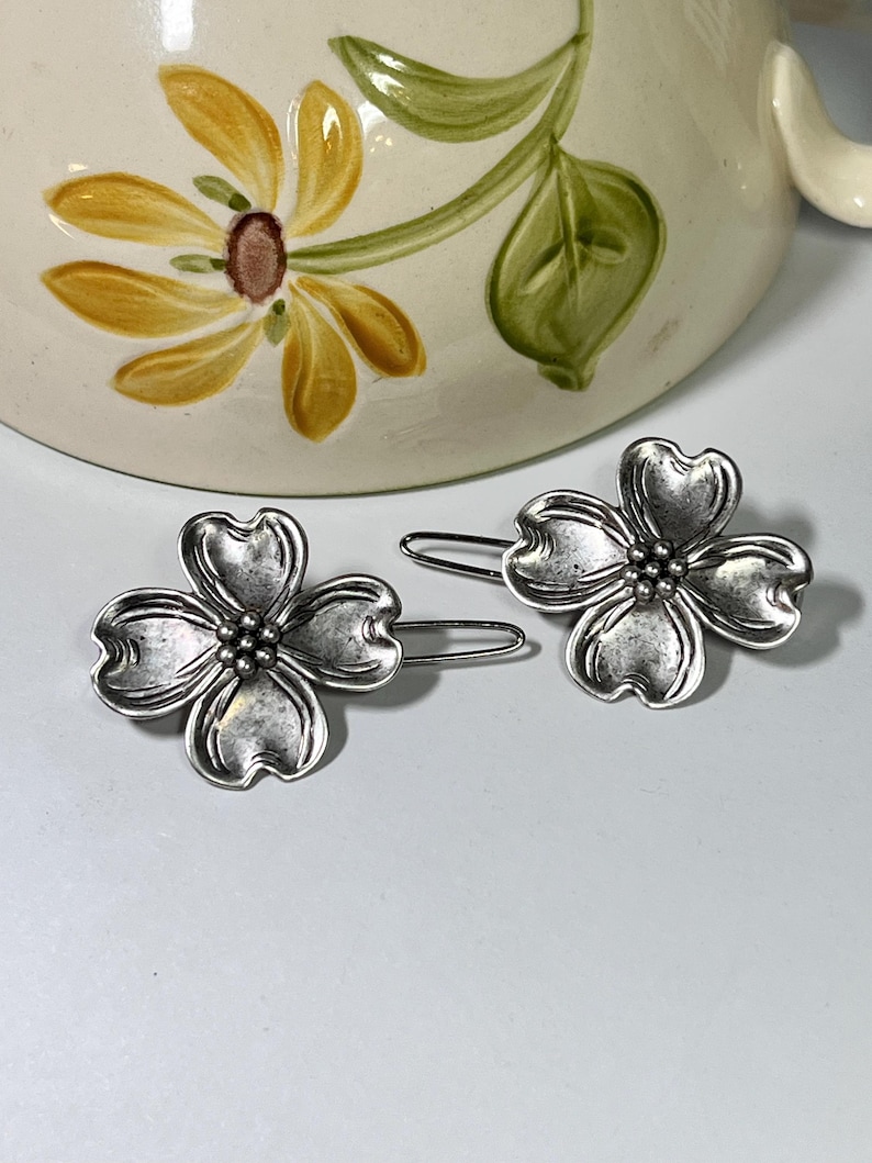 May include: Two silver-toned flower-shaped hair clips. Each clip features four petals and a cluster of small beads at the center. The clips are set against a light background, with a floral-decorated ceramic mug in the background.