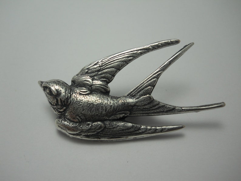 May include: A detailed silver-colored bird brooch in flight, with outstretched wings and detailed feathering. The bird's beak is slightly open, and its tail feathers extend backward. The brooch is likely made of metal and has a textured surface.