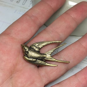 May include: A small, antique gold-colored swallow bird charm. The bird is in mid-flight with detailed wing and tail feathers. The charm has a vintage, weathered appearance, suggesting it is made of metal.