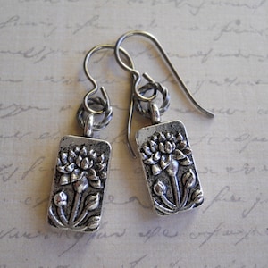 May include: Silver earrings with a delicate floral design. Each earring features a rectangular charm with a raised image of a flower.