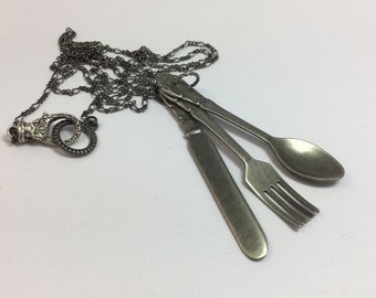 Może przedstawiać: Srebrny naszyjnik z zawieszką przedstawiającą nóż, widelec i łyżkę. Łańcuszek wykonany jest z małych, połączonych kółek.