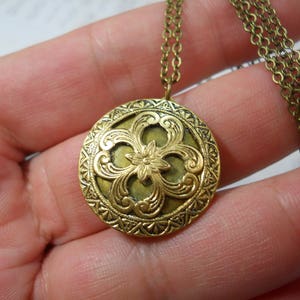 May include: A round, antique brass-colored pendant necklace. The pendant features an ornate floral design with a central flower and swirling patterns. The necklace has a delicate chain.