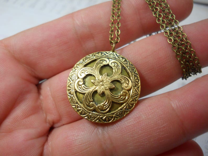 May include: A round, antique brass-colored pendant necklace. The pendant features an ornate floral design with a central flower and swirling patterns. The necklace has a delicate chain.