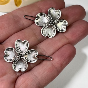 May include: Two silver-toned hair barrettes, each shaped like a dogwood flower. The barrettes have a detailed, textured design with a cluster of small spheres at the center of each flower. The barrettes are on a silver-colored metal clip.