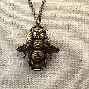 May include: A gold-toned necklace featuring a bee-shaped locket pendant. The locket is detailed with a textured bee design and has a hinged opening.
