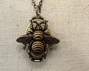 May include: A gold-toned necklace with a bee-shaped locket pendant. The locket is detailed with a textured bee design and has a hinged opening.