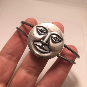 May include: A silver bracelet with a round face featuring a smiling man's face. The bracelet is made of two thin metal bands.