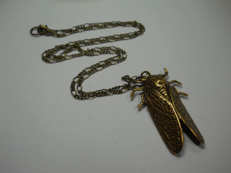 Puede incluir: Un collar dorado con un colgante de cigarra detallado. La cadena es de estilo de doble enlace.