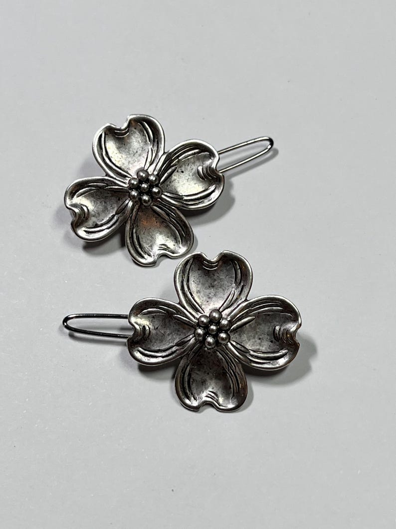 May include: Two silver-toned metal hair clips. Each clip features a flower design with four petals and a cluster of small beads at the center. The clips have a simple, curved bar for insertion into the hair.