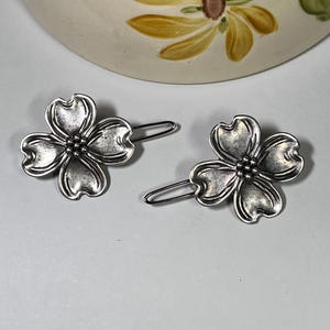 May include: Two silver-toned metal hair clips, each shaped like a four-petal flower. The clips have a detailed, textured surface and a simple, curved pin backing. The background includes a portion of a ceramic item with a floral design.