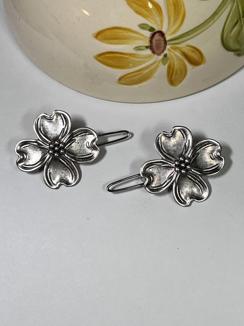May include: Two silver-toned metal hair clips, each shaped like a four-petal flower. The clips have a detailed, textured surface and a simple, curved pin backing. The background includes a portion of a ceramic item with a floral design.