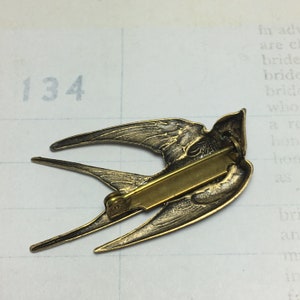 May include: A vintage-style bronze bird pin with outstretched wings. The pin features intricate detailing of the bird's feathers and body. The pin has a gold-colored clasp on the back. The background is a vintage paper with the number 134.