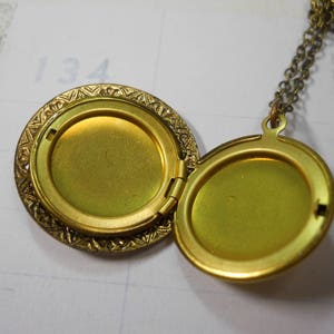 May include: Open, antique gold-colored locket necklace. The round locket is hinged and opens to reveal two compartments. The locket has an ornate, textured border and hangs from a delicate chain. The background is a light-colored paper.