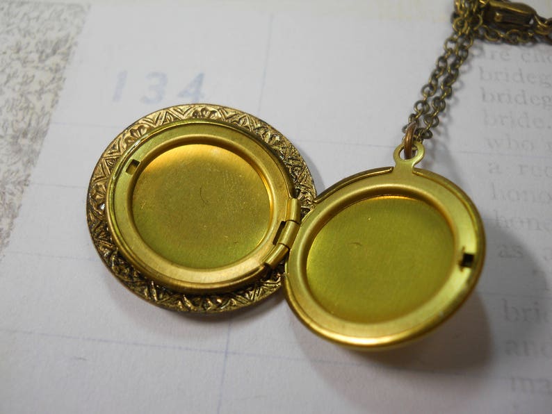 May include: Open, antique gold-colored locket necklace. The round locket is hinged and opens to reveal two compartments. The locket has an ornate, textured border and hangs from a delicate chain. The background is a light-colored paper.