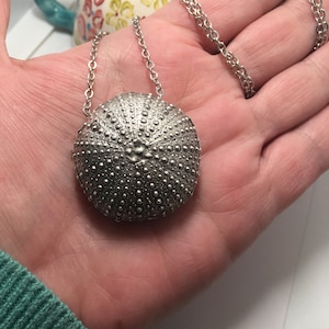 May include: A silver necklace with a round pendant that resembles a sea urchin. The pendant is detailed with small, raised dots.