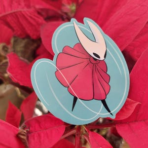 May include: A sticker featuring a white and red character with a flowing red cape and a blue background. The character is a popular video game character.