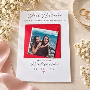 May include: A white card featuring the text Dear Natalie, I can't imagine my wedding day without you, Will you be my bridesmaid?, and the date 23 08 2026 with a small pink heart. A square photo of two people is attached over a red envelope.
