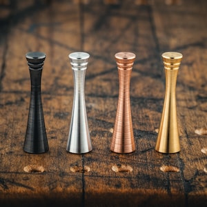 May include: Four metal bottle stoppers in a row, each with a unique finish. The stoppers are black, silver, copper, and gold. They have a flared, conical shape with a cylindrical top. The stoppers are displayed on a wooden surface.