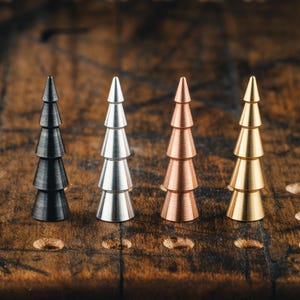 Peut inclure: Quatre ornements métalliques en forme de sapin de Noël, de couleurs noir, argent, cuivre et or. Chaque arbre a une forme conique avec des couches superposées, posé sur une surface en bois. Les ornements sont disposés en rangée, mettant en valeur leurs différentes couleurs.