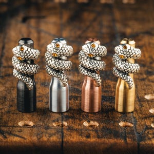 Puede incluir: Cuatro tapones de botella de metal con diseños de serpiente. Cada tapón presenta una serpiente enroscada con ojos de color ámbar. Los tapones son de color negro, plateado, cobre y dorado. El fondo es una superficie de madera.