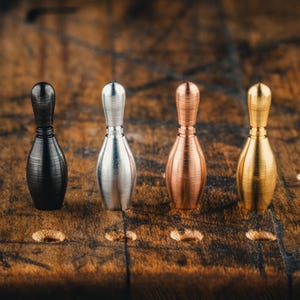 Peut inclure: Quatre sculptures miniatures en forme de quilles de bowling, de couleurs noire, argentée, cuivrée et dorée, sont présentées sur une surface en bois texturé. Les quilles ont une finition lisse et métallique, de forme classique.