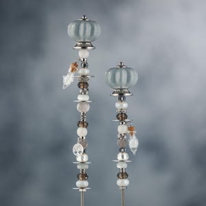 May include: Two decorative metal garden stakes with a silver finish. Each stake features a white ceramic knob with a gray and white striped pattern, and is adorned with a variety of beads, charms, and glass vials.
