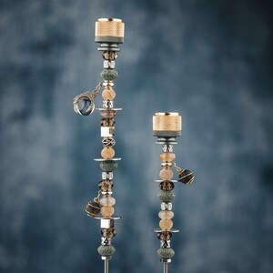 May include: Two decorative metal rods topped with textured gold-toned cylindrical caps. The rods are adorned with a vertical arrangement of beads in shades of gray, brown, and gold, interspersed with silver-toned spacers and small dangling charms.