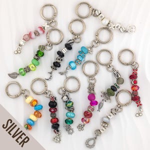 May include: A collection of ten silver-toned metal keychains, each featuring a circular ring and a unique arrangement of colorful beads, charms, and decorative spacers. The charms include shapes like butterflies, moons, crabs, fish, and cherries.