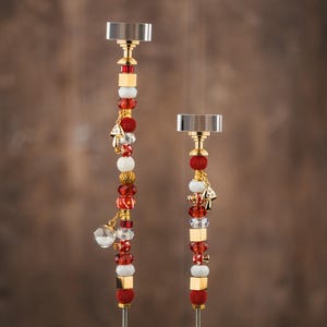 May include: Two decorative candle holders with silver tops and gold accents. The stems are adorned with red, white, and gold beads, along with small charms. The candle holders are set against a brown background.