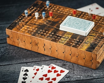 May include: A wooden cribbage board with a brown finish and a yellow and black checkerboard pattern. The board is made of multiple pieces that fit together. There are four playing cards on the board, including a 9 of spades, a 9 of diamonds, an ace of spades, and a 10 of hearts. There are also several small red and blue pegs on the board.