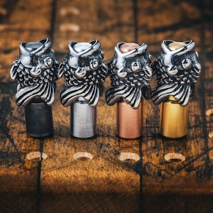 May include: Four metal bottle stoppers with owl-shaped tops in silver, black, copper, and gold. The stoppers are on a wooden surface, showcasing the detailed craftsmanship of the owl design. The stoppers are designed to fit standard bottle openings.