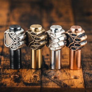 Könnte beinhalten: Vier Flaschenverschlüsse aus Metall mit Pfotenabdruck-Design. Die Verschlüsse sind in Schwarz, Messing, Silber und Kupfer.