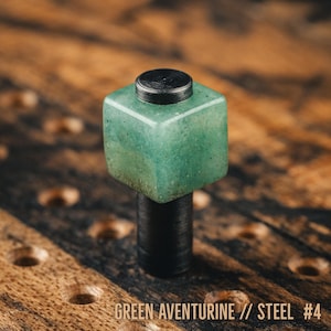 May include: A green aventurine and steel bottle stopper. The stopper features a square, light green aventurine top with a black steel base. The text "GREEN AVENTURINE // STEEL #4" is at the bottom.