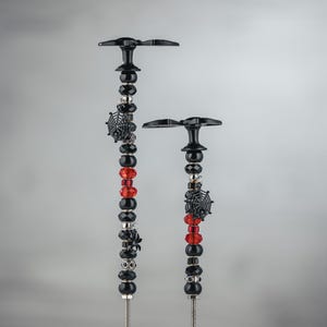 Puede incluir: Dos estacas decorativas para jardín con adornos de cuentas negras y plateadas. Las estacas presentan dijes de telaraña negra, cuentas rojas y negras y detalles plateados. Las estacas tienen una parte superior negra en forma de mariposa.