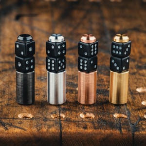 Könnte beinhalten: Vier Flaschenverschlüsse in Würfelform mit schwarzen Würfeln und metallischen Basen in Schwarz, Silber, Kupfer und Gold. Die Würfel sind gestapelt und haben weiße Punkte. Die Verschlüsse werden auf einer Holzoberfläche präsentiert.