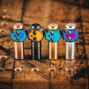 Op de afbeelding: Vier metalen flessenstoppers in de vorm van een schedel in verschillende afwerkingen: koper, zwart, goud en zilver. Elke stopper heeft een regenboogkleurige, iriserende afwerking.