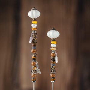 May include: Two decorative garden stakes with white ceramic tops and brown, yellow, and silver beads. The stakes are made of metal and have a rustic, bohemian style.