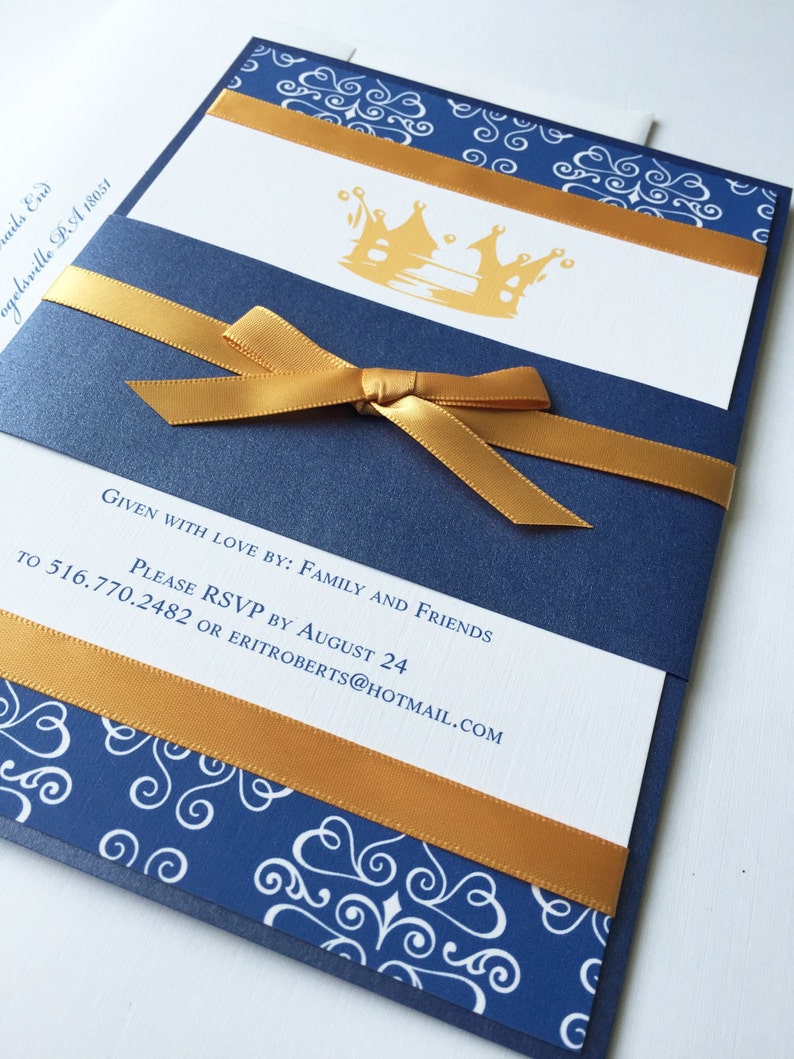 Op de afbeelding: Een blauwe en witte uitnodiging met een gouden lint en een gouden kroonontwerp. De tekst op de uitnodiging luidt "Given with love by: Family and Friends. Please RSVP by August 24 to 516.770.2482 or eriroberts@hotmail.com"