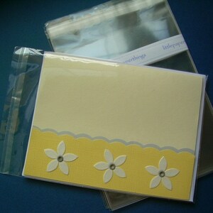 May include: A set of greeting cards in clear cellophane packaging. The cards have a light yellow background with a yellow textured border and white flower embellishments. The text "littlepaperthings" is visible on the packaging.