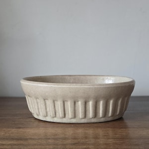 May include: A beige, oval-shaped ceramic baking dish with vertical ribbed detailing around the sides. The dish sits on a wooden surface against a neutral background. This kitchenware item is suitable for baking and serving.
