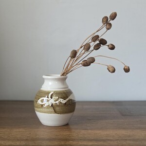 May include: A small, ceramic vase with a white top and base, and a brown and white patterned middle section. The vase holds several dried, brown seed heads on slender stems. The vase sits on a wooden surface.