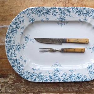 May include: An oval white ceramic serving platter featuring a blue floral pattern along the rim, accompanied by a metal knife and a three-tined fork, both of which have light-colored, riveted handles positioned horizontally in the center.