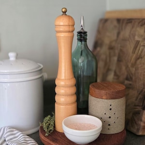 May include: A collection of kitchen items on a wooden surface. Includes a tall wooden pepper grinder, a green glass bottle, a small white bowl with pink salt, and a ceramic container with a wooden lid.