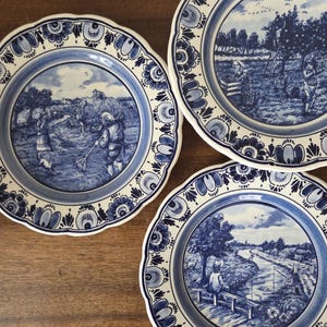 May include: Three decorative ceramic plates with blue and white pastoral scenes. Each plate features a scalloped edge with floral and geometric patterns. The scenes depict people working in fields and near water.