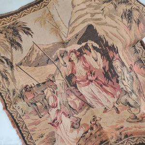 May include: A vintage tapestry depicting a scene of people and animals in a desert setting. The tapestry features a brown, beige, and pink color scheme and includes a camel, a dog, and several people in traditional clothing.