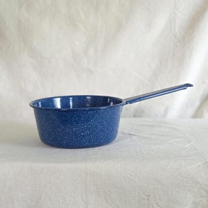 May include: A blue speckled enamel saucepan with a long handle. The pan is round with a slightly flared rim. The handle is attached to the side of the pan and is the same blue color. The pan is sitting on a white surface.